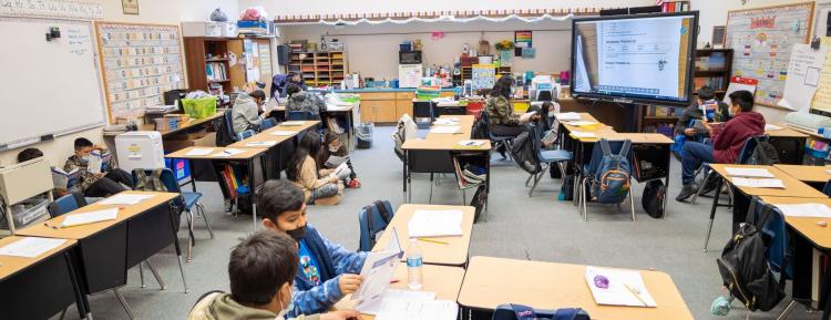 A classroom with students sitting at their desks, and a projector screen where a teacher teaches.