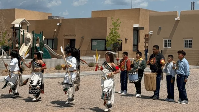 Several children in traditional Pueblo attire are dancing while holding feathers and other ceremonial items.