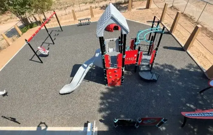 Aerial view of a small outdoor playground on rubberized black surfacing, featuring a red-and-gray play structure with a slide, climbing wall and overhead bars.