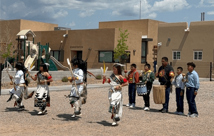 Several children in traditional Pueblo attire are dancing while holding feathers and other ceremonial items.