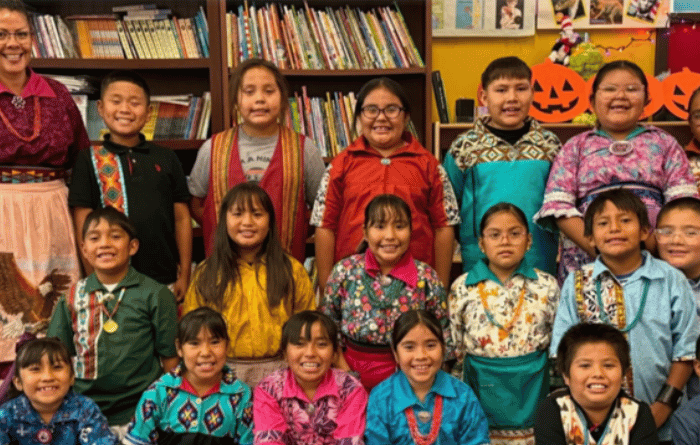 A group of children and a teacher smile for a photo in a school library, wearing colorful clothing and standing in front of bookshelves and Halloween decorations.