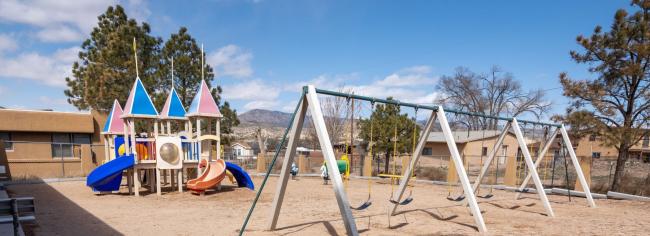 A playground featuring swings and slides set against a backdrop of a vast desert landscape under a clear blue sky.