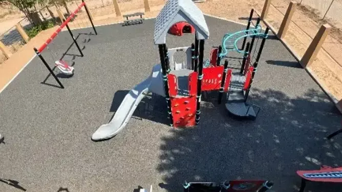 Aerial view of a small outdoor playground on rubberized black surfacing, featuring a red-and-gray play structure with a slide, climbing wall and overhead bars.
