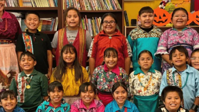 A group of children and a teacher smile for a photo in a school library, wearing colorful clothing and standing in front of bookshelves and Halloween decorations.